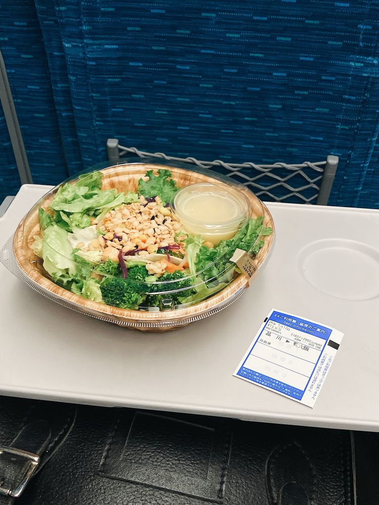 新幹線の座席のテーブルに、テイクアウトしたサラダと、品川から新大阪のチケットが置かれている。