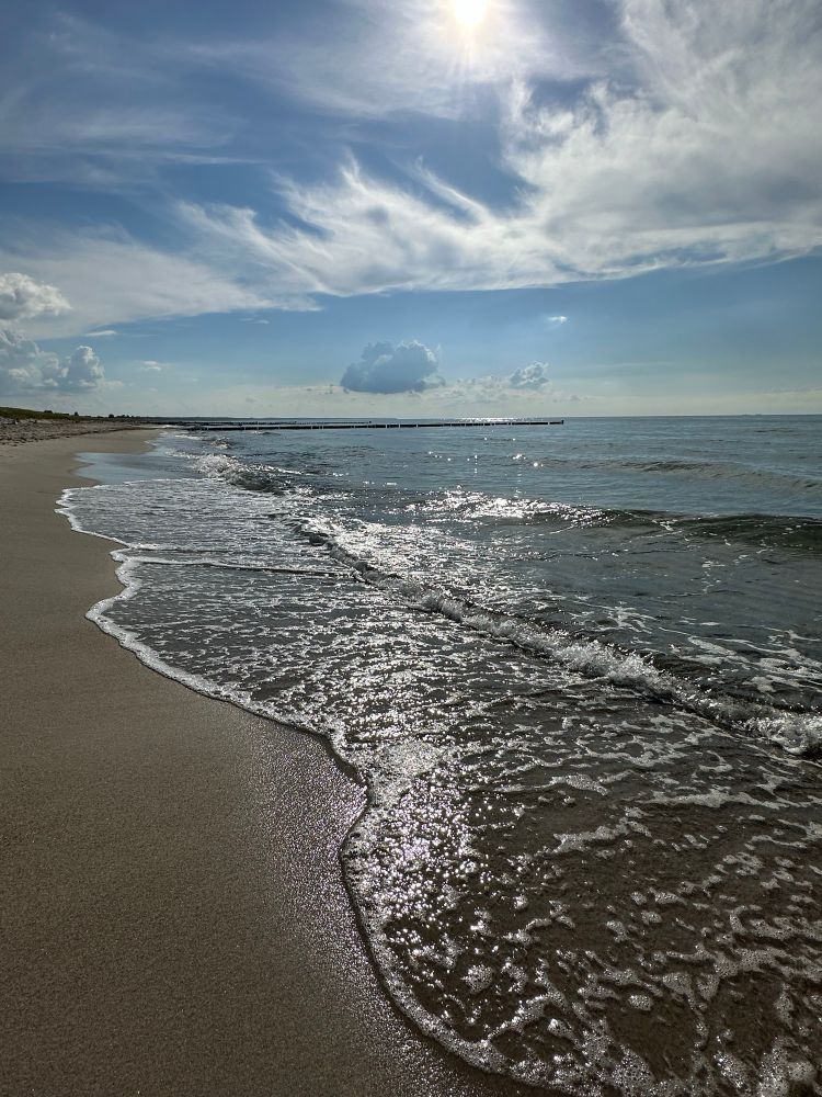 Die Sonne scheint auf die Meeresbrandung und den Sandstrand und leuchtet sie hell an. Der Himmel am Horizont ist blau mit ein paar Federwolken und ein paar kleineren Haufenwolken. 
