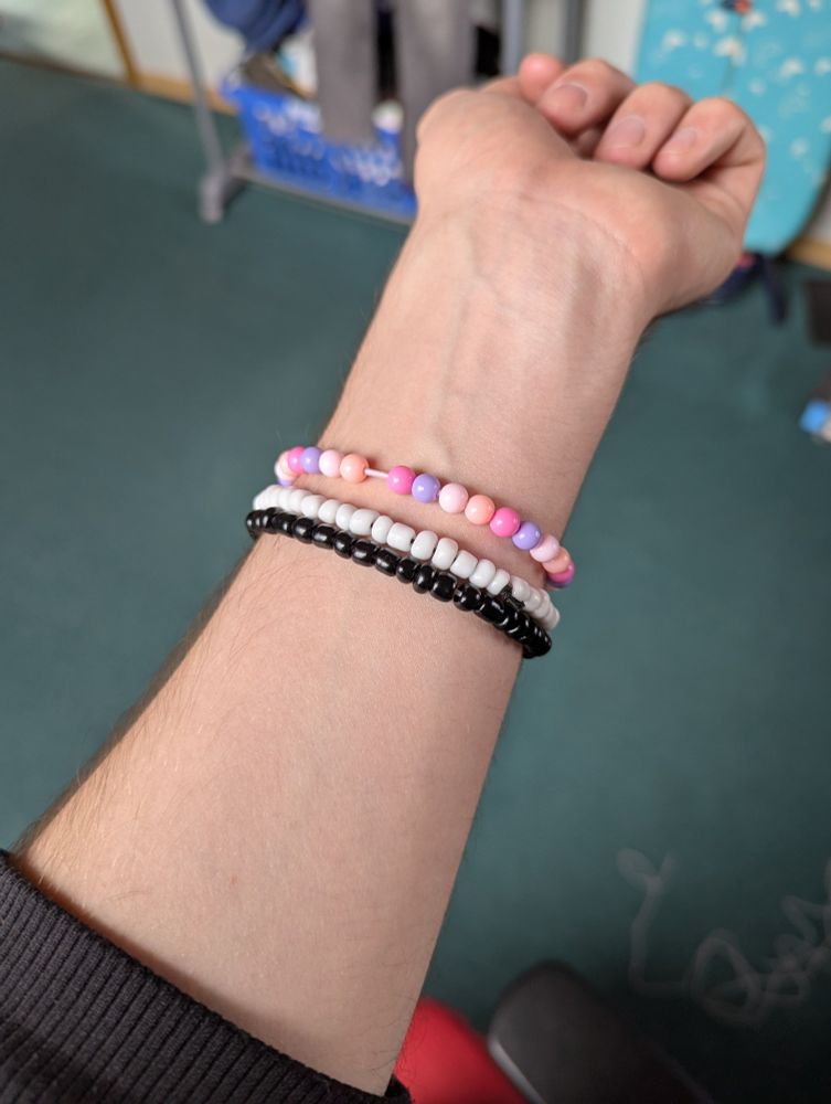 my arm with three self-made bead bracelets on, the first with round plastic beads in pastel pink/purple colors, the other two with irregularly-shaped ceramic beads – one white, one black