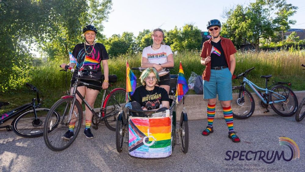 Four people on a paved road, standing or sitting with bicycles, holding or displaying Pride flags. The person on the left is standing beside a black road bike, wearing a black t-shirt with a rainbow flag design, black shorts, and rainbow-striped socks. The person in the center front is sitting in a three-wheeled adaptive bicycle with a rainbow flag design on the front panel, wearing a black t-shirt and glasses. Behind them, another person stands beside a bicycle, wearing a white t-shirt that says "love is love". On the right, a person stands wearing a red shirt, teal shorts, rainbow-striped socks, and holding a small Pride flag. Two more bicycles are parked in the background on the right. The sun shines brightly on the trees and foliage in the background. In the bottom right corner is a logo for "SPECTRUM Waterloo Region's Rainbow Community Space".