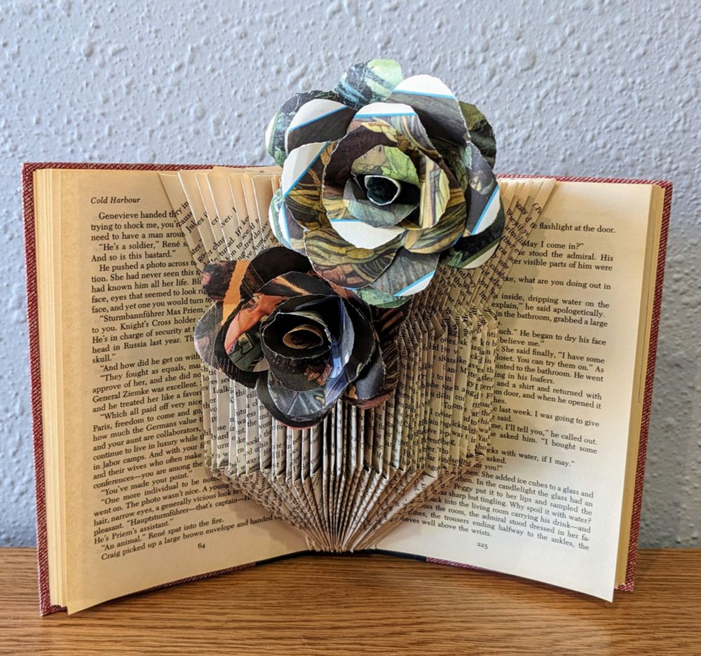 A thin Readers Digest condensed book (about 3/4 inch thick) with the center 160 pages folded into the shape of a vase with a narrow base, wide center, and flared top. There are two paper roses, one at the middle and slightly left and the other at the top and slightly right. The lower rose is in shades of black, gray,  and orange. The top rose is in shades of white, yellow, and green.