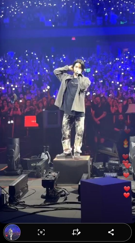 Yoongi stands in a spotlight, on a black box on a black stage, holding a mic to his mouth with 1 hand, the other is maybe scratching the back of his head. Behind him, an audience is packed in, drenched with red light on the front rows, purple light for the next few rows, and then light blue toward the back. There are dots of white light in the audience, either phone flashlights or ARMY bomms.
