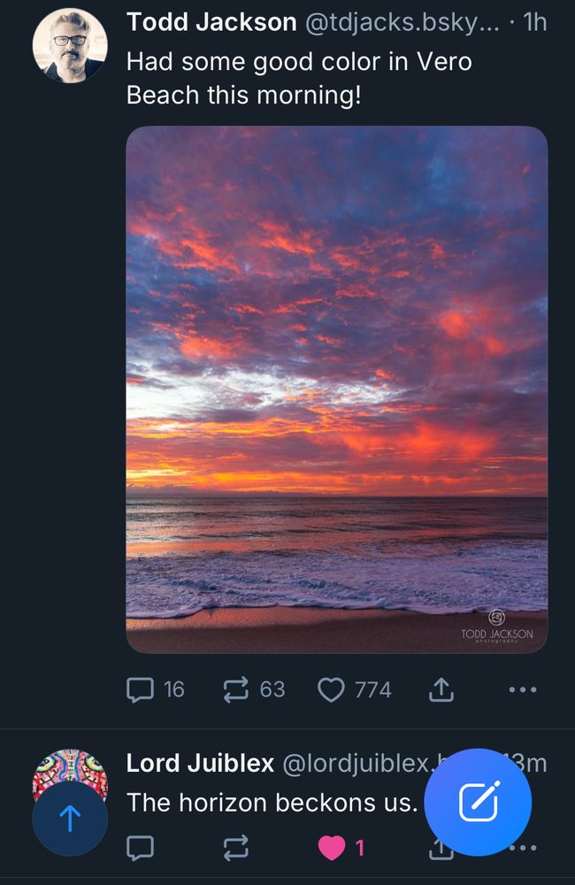 A photo of the horizon over the ocean, the sun is rising and soft, fluffy clouds are piled up in bunches overhead, a dazzling array of crimson, gold, and indigo light playing across their surface. 
