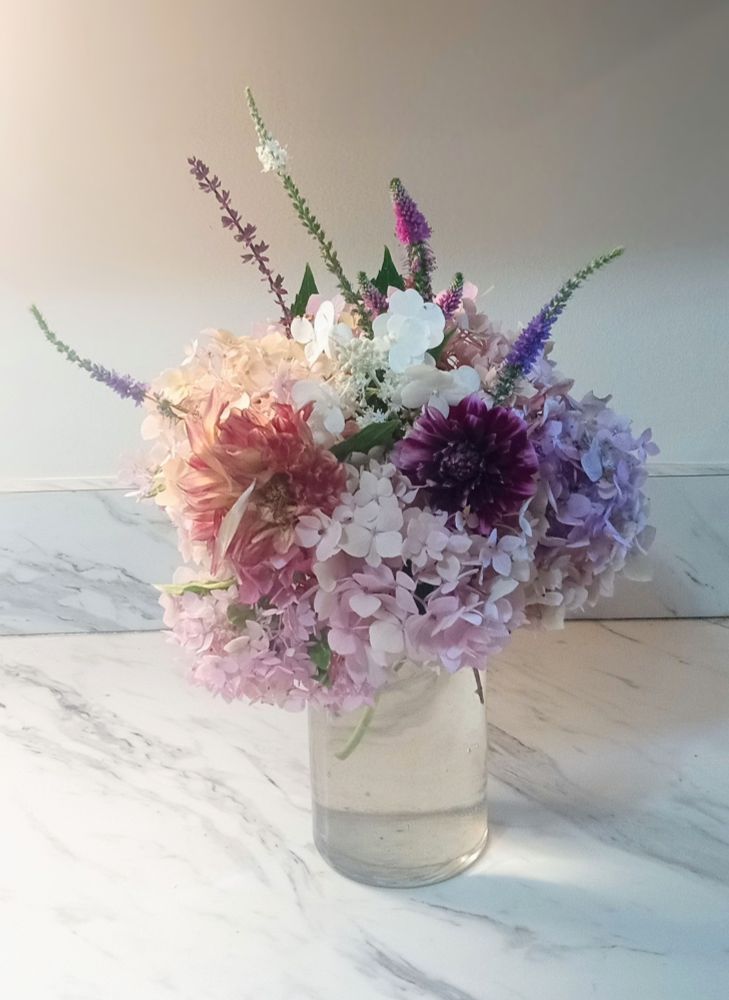 A picture of an assortment of flowers in a clear vase on a counter. They're Hydrangeas, Dahlias, Veronica, and sage.
