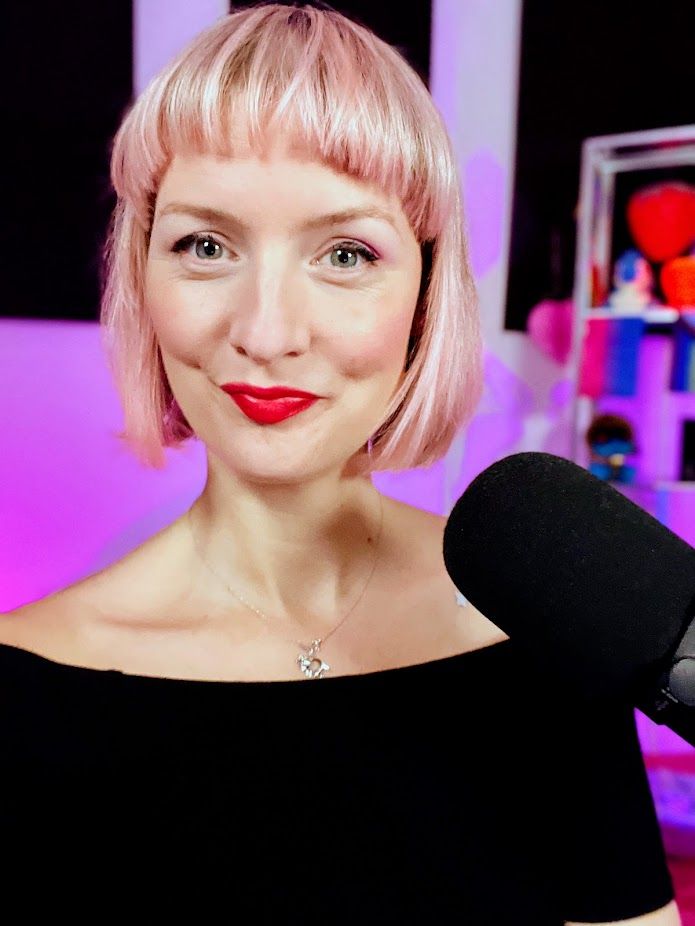 A brightly-lit selfie of MollyBerry, taken at her streaming setup in her studio. Her hair is pastel pink/blonde and she's smiling.
