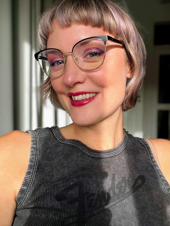 A selfie of MollyBerry, smiling as she escapes the sun streaming through the windows of her home in Lisbon, PT. She's wearing glasses, and has a short, cropped fringe and a French-style bob haircut. Her hair is shades of pastel purple, blonde, platinum, and grey. Molly is wearing a grey tank top with the brand logo of Fender across the chest. Photo was taken November 3rd, 2025.