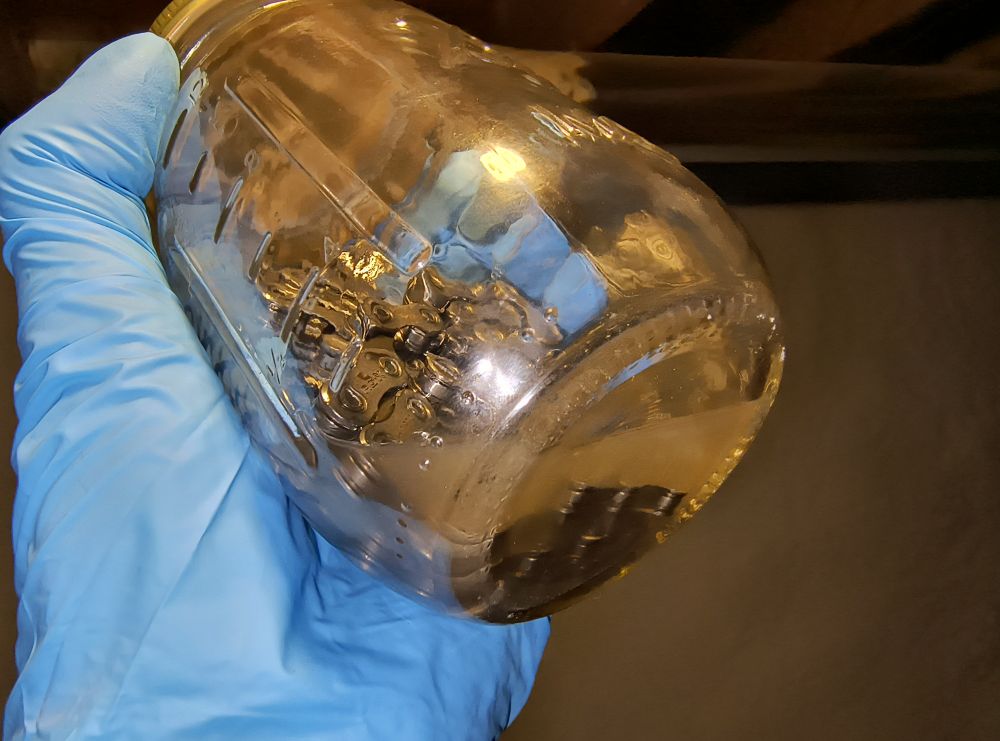 Color photo of a small glass mason jar held by a nitrile gloves hand. Within the jar is 2oz of Finish Line Citrus degreaser and a length of Shimano HG-X chain