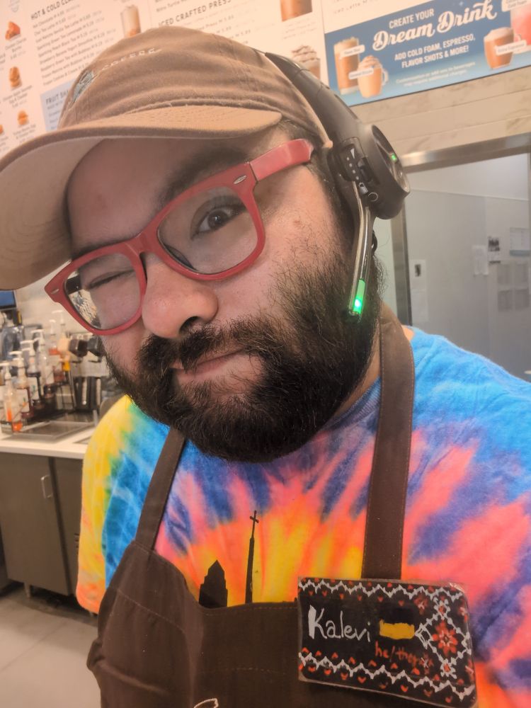 Demi guy winking into the camera, wearing a drive thru headset, a brown apron with a cut-off corporate logo, a name tag that says "Kalevi + he/they" and is designed to look like vyshyvanka embroidery from Kharkivshchyna, and also displays a small Ukrainian flag. Also wearing a rainbow tie dye T-shirt that has a covered up church logo. (Ordered for the Pride parade!)