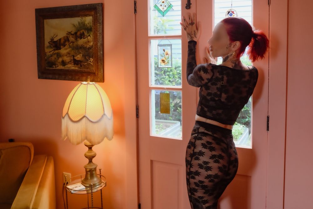 Magnolia wears a long sleeved black lace top with see through lace pants. Her red hair is pulled back and she stands by the door with both hands on the wall, back arched. 