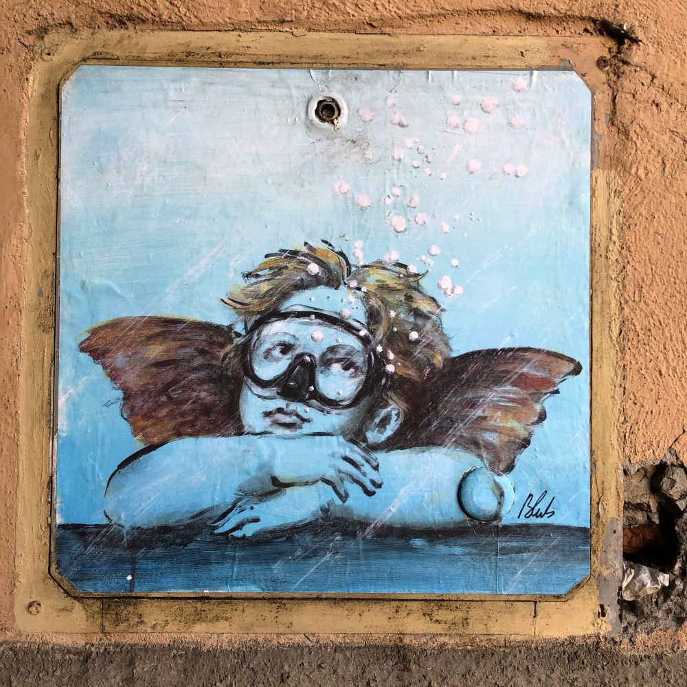 Street art - winged cherub with blond hair and swim goggles, head resting on arms.