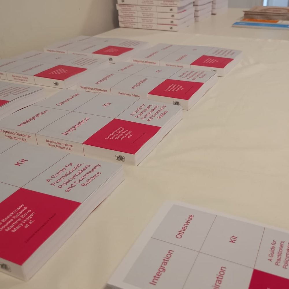 A table is neatly arranged with multiple copies of the Integration Otherwise Inspiration Kit, a book with a clean white and red cover design. The subtitle reads: “A Guide for Practitioners, Policymakers, and Community Builders.” The authors’ names—Beeckmans, Salamé, Bovo, Hogan et al.—are visible on the covers. The books are stacked and spread out, ready for distribution at the event.