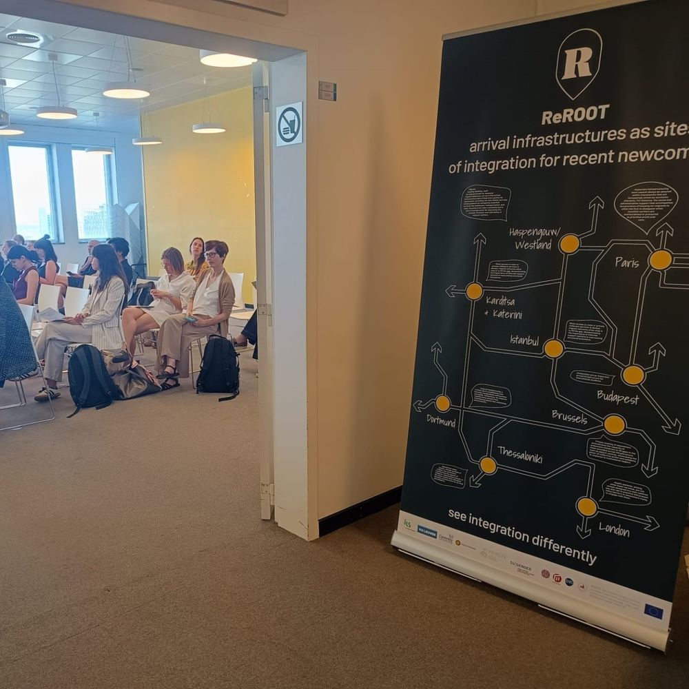 A ReROOT project banner stands at the entrance of a conference room, showing a stylized network map with cities like Brussels, Paris, Thessaloniki, and Dortmund connected by arrows and speech bubbles. The title reads “ReROOT: arrival infrastructures as sites of integration for recent newcomers” with the slogan “see integration differently” at the bottom. Inside the room, participants are seated and listening attentively, with natural light streaming in through large windows.