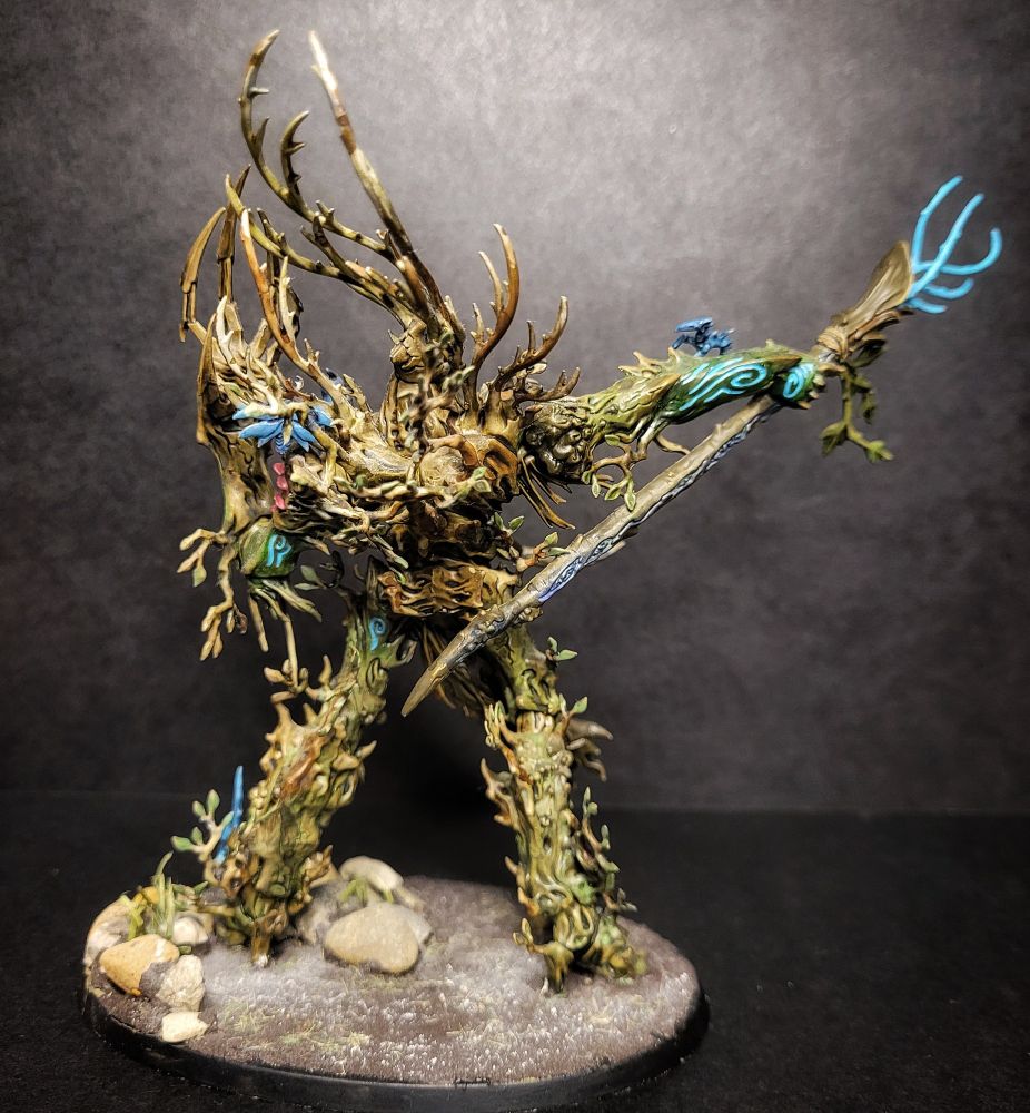Age of sigmar tree lord from the back covered in tiny indigo woodland sprites. The tree lord is painted in a warm green brown colour with blue / turquoise accents, in this picture you can see blue tendrils coming out if it's staff.