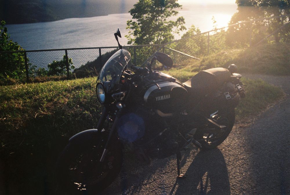 A motorcycle viewed sideways, with far below a Norwegian fjord. The sun shines almost directly at the camera, just out of frame to the top-right. 