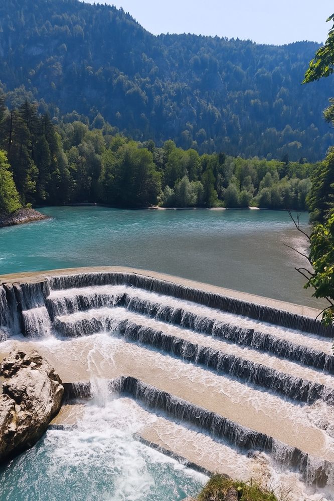 The Lechfall in Füssen: a stepped artificial waterfall where the turquoise river Lech cascades down several wide stone ledges. Trees line the banks, and the flow continues into the valley beyond.
