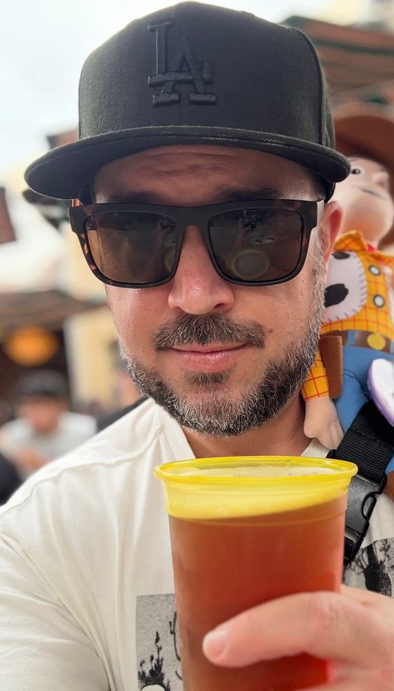 A selfie of me at Disney’s California Adventure with sunglasses, an old Silly Symphonies shirt, and a black Dodgers hat, drinking a Red Trolley Ale from Karl Strauss, with a Woody figure on my shoulder that I won at Pixar Pier.