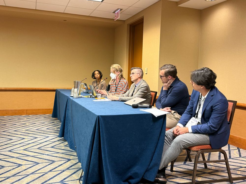 Presenters discuss their dissertation research seated at a long table in a conference room