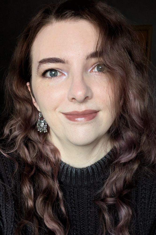 Tulip smiling, wearing a dark grey sweater, silver dangling earrings, and a touch of iridescent blue eyeshadow on their eyes. they have long, purple toned brown hair