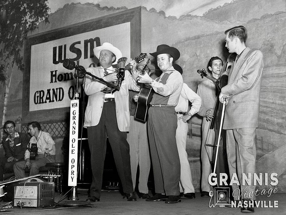 A horizontal black and white film photo of Bill Monroe, Jimmy Martin, Buddy Killen, and Don Slayman playing instruments on stage with Chet Atkins watching on stage behind them