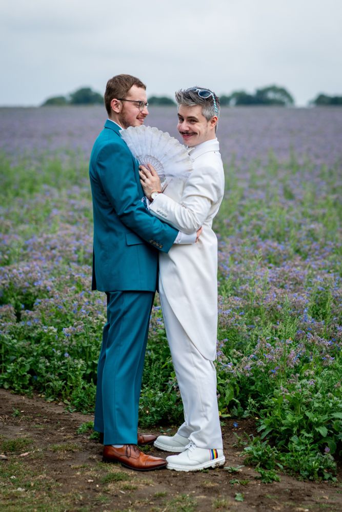 Dan and Arthur standing in a field of lavender in their wedding suits, Dan is holding a fan and looking at the camera, Arthur is looking at Dan