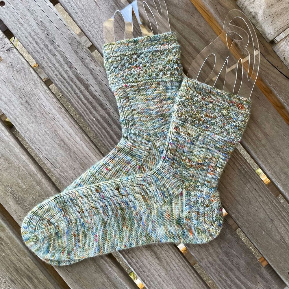 A pair of hand-knit socks on clear acrylic blockers, displayed on a wooden chair.  The yarn is softly variegated in shades of blue, green, and tan.  The pattern is mostly ribbed, but features a slip-stitch textured band on the cuff.  
Pattern:  Ensnared by Hunter Hammersen.
Yarn:  Swift Yarns Bliss Sock in Antique Sky.