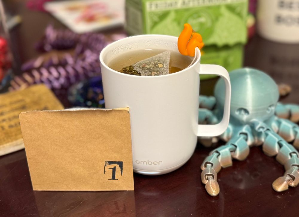 A torn-open brown packet labeled 1 sits propped against a white cup of brewing tea.  A pale blue 3D printed octopus sits nearby, guarding against any would-be tea thieves.  