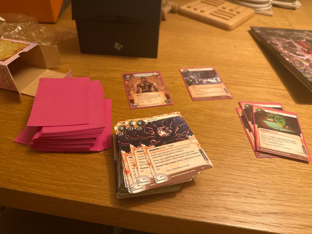A stack of Netrunner cards (the runner side) and the pink diamond sleeves I’ve chosen to be the runner color. All of this is against a backdrop of a wooden table, with the following in the background:

* an open box that previously housed the sleeves (and the plastic wrap that enclosed the box)
* the black storage box I’m using as a card storage solution for the time being. Eventually I will use something bigger but I’m not ready yet.
* the corner of a Pathfinder 2E book, Rival Academies