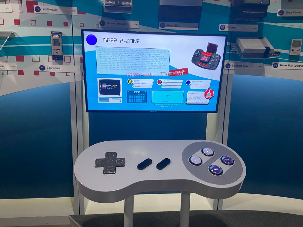 A television screen with a giant Super Nintendo controller in front of it. The screen is displaying information about the Tiger R-Zone console. The controller can be used to navigate through a menu of various other consoles and bring up information about each of them. On the wall behind the screen, several of the consoles are mounted on display, including an NES and an original Game Boy