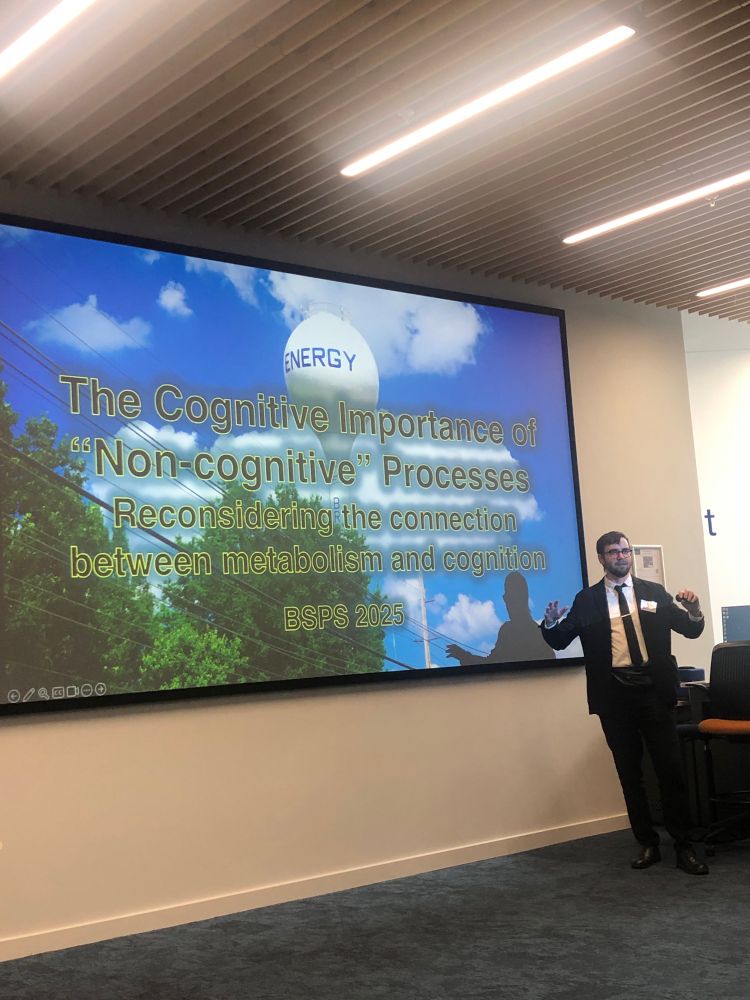 Philosopher David Colaço, eating a black suit jacket, white shirt and black tie in front of an image with a white balloon with „energy“ written in blue, a blue sky and white clouds on the back, some green tree canopies in front. 