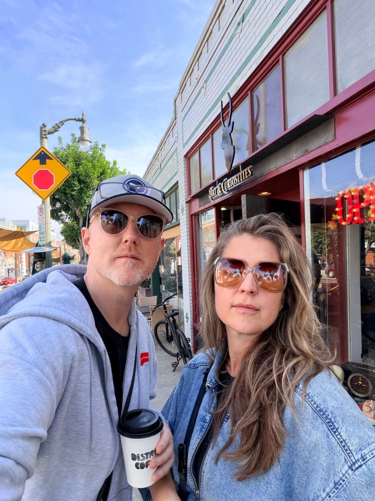 Selfie of me in a hat and glasses holding a coffee cup with a blonde woman also in sunglasses