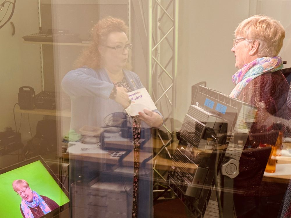 Zwei Frauen sprechen miteinander in einem Fernsehstudio. Eine Frau steht vor dem Greenscreen und spricht zur Kamera. Die andere Frau hält einen Zettel mit Text und scheint der Frau vor der Kamera Hinweise zur Aussprache zu geben.