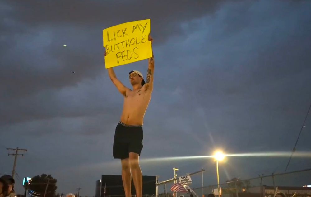 A shirtless man holds aloft a yellow sign in the twilight outside a Chicago ICE facility. Its message, handwritten in black sharpie: "lick my butthole feds"