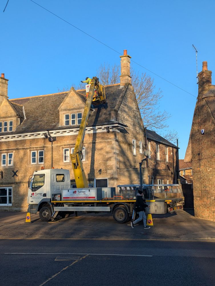 Two men wearing high visibility clothing standing on a hydraulic cherry picker maintaining a roof of a public house in Peterborough, the weather sunny with blue skies