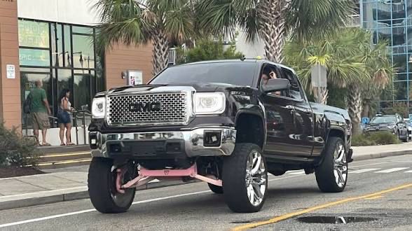A large black GMC crew cab pickup with a now-banned “Carolina Squat” mod, in which the front axle is dramatically jacked up, but the back is not, so the driver has horrendous front visibility but at least it looks idiotic and was extremely expensive