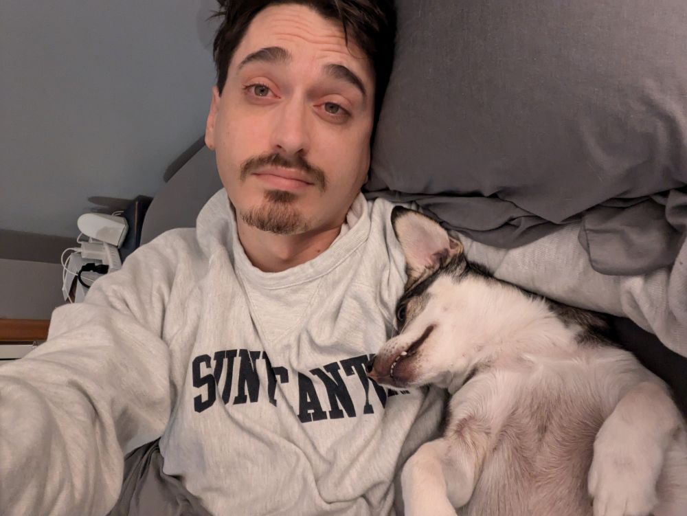 A man with short dark hair and a goatee is lying on a bed wearing a light gray sweatshirt that reads "SUNNY CANTON." Beside him is a small dog lying on its back, showing its belly with a slightly mischievous grin. The man has a calm expression, and the atmosphere is cozy.