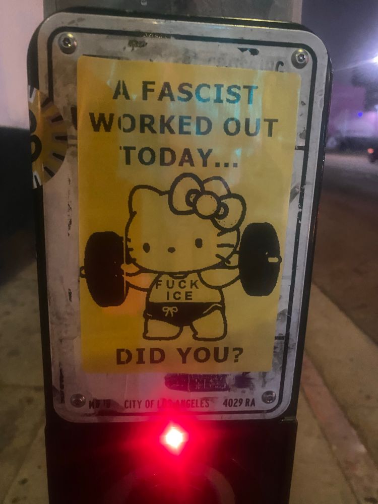 A sticker placed at a walk signal for a stop sign. In black on yellow paper, a Hello Kitty is shown lifting weights in a tank top. Text on the shirt reads, “FUCK ICE.” Text on the sign reads, “A fascist worked out today… did you?”