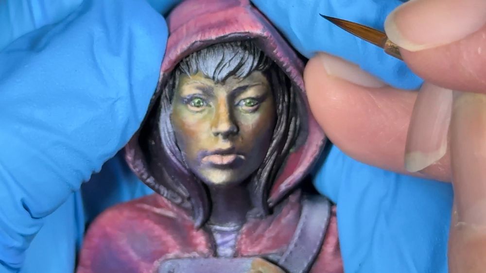 Bust (Lillith, by Bold Miniatures), halfway through the painting process, zoomed in on the face; she's wearing a purple-red hood, and there's green, orange, and purple tones painted onto the face for mood/temperature sketching (which ended up being the final color since I loved it). Her eyes are bright green, and probably the best eyes I've painted so far.