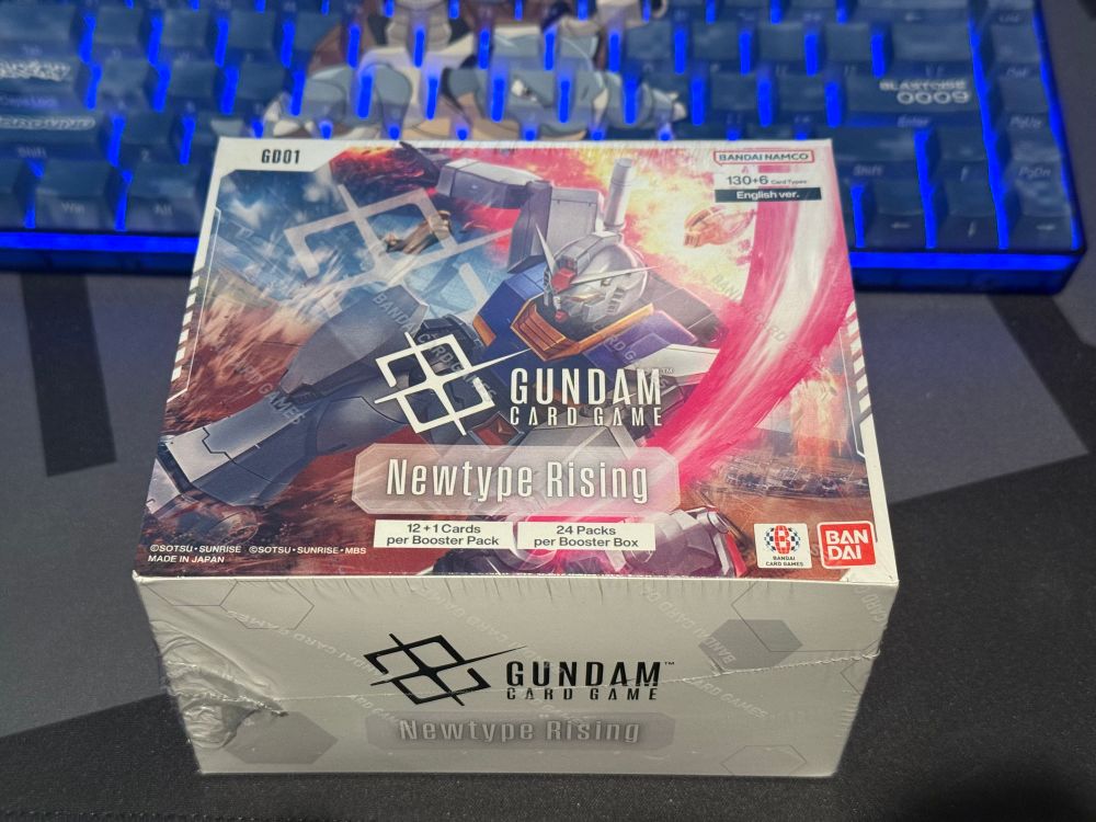 An unopened box of Gundam Card Game: Newtype Rising booster packs on a grey and black mat, in front of a Blastoise keyboard 