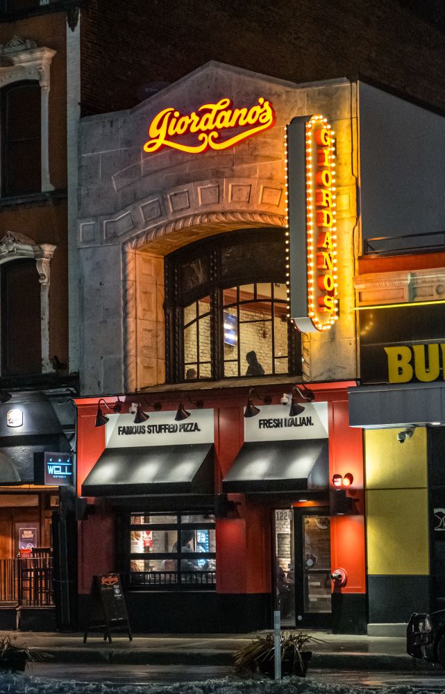 Color photograph taken at night of a lighted two story restaurant in downtown Detroit. And an upper arched and planed window you can see the silhouette of someone sitting.