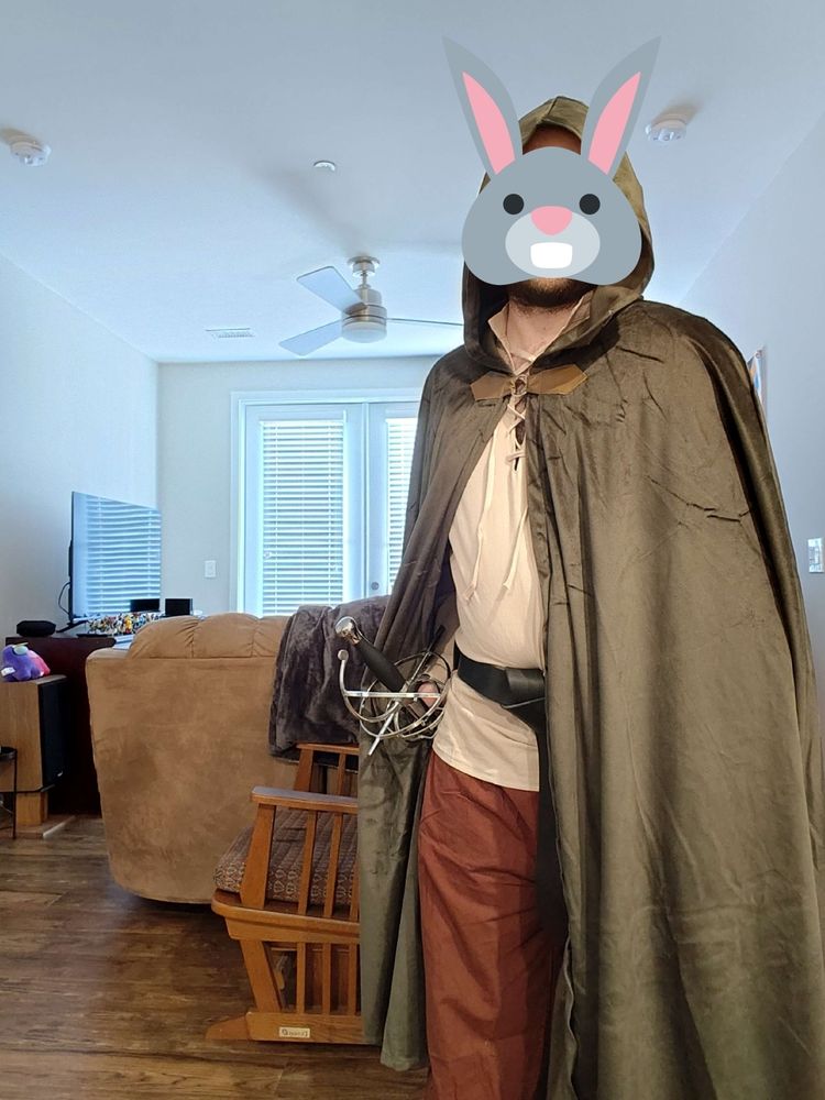 A man with a rabbit emoji obscuring his face, wearing a cloak with the hood drawn, one half held to the side to show his rapier