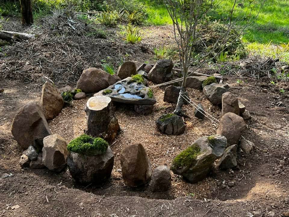 Picture of a circle made of stones.  Inside is a large flat stone with hollows filled with water, a small dogwood tree, and a stump for sitting on. Some of the stones have moss on them. Pacific Northwest. 