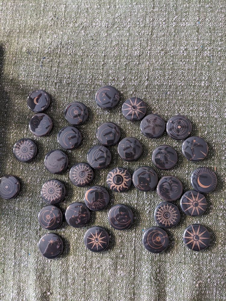 A group of about two and a half dozen buttons sit on a table. They are all of various golden designs on black backgrounds, mostly featuring celestial motifs such as moons, stars, suns, and cryptic eyeballs.