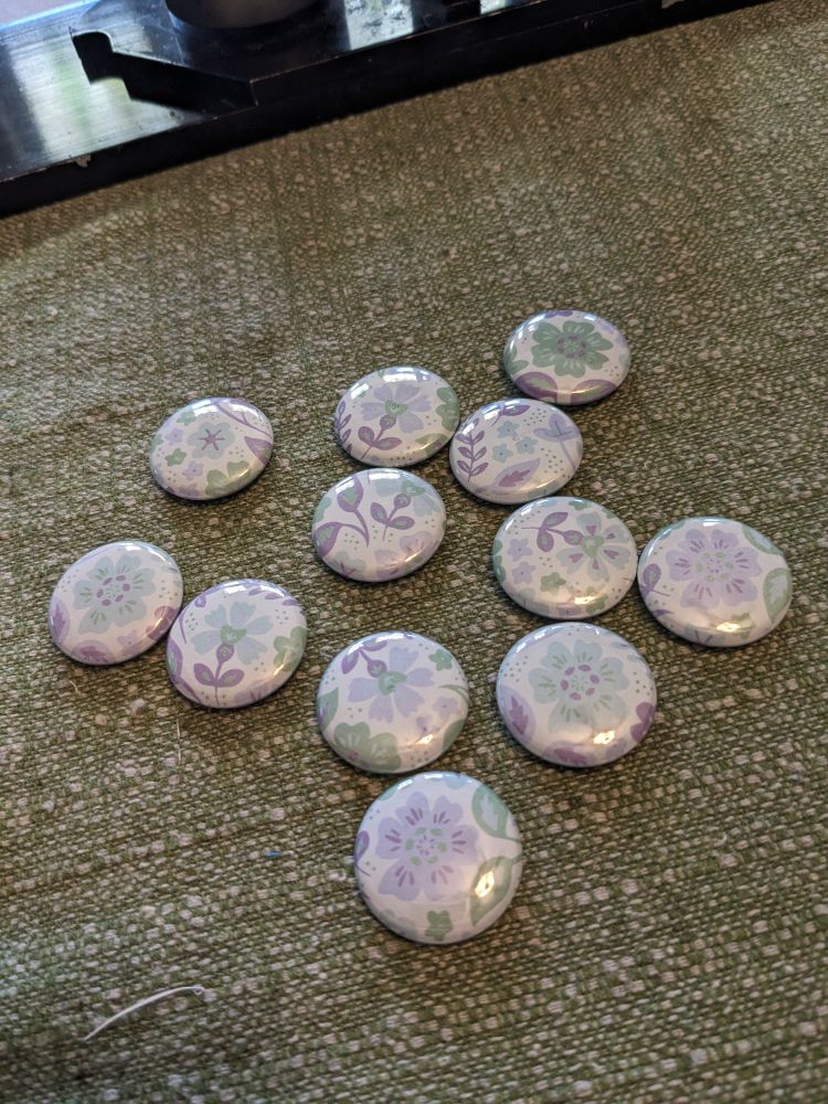 A group of about a dozen buttons sits on a table. The buttons are all floral designs on white backgrounds, colored with green and lavender hues.