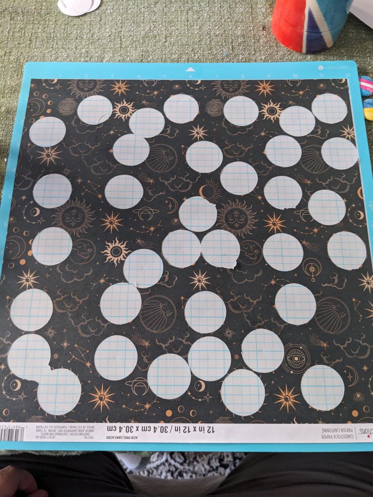 A sheet of patterned paper sits on an adhesive cutting mat with many circles cut out of it. The pattern is celestial motifs in gold on a black background, including moons, stars, suns, and other zodiac-inspired shapes.