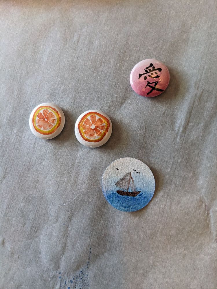 A small selection of water color painted button designs. One is a pink gradient background with the sinograph 愛, two are orange slices, and one is a sail boat at sea, still waiting to be pressed into a button.