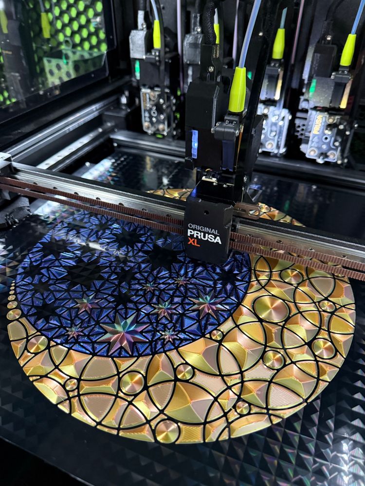 In progress shot of a Prusa XL 3D printer producing a 3D printed mosaic circle in shimmering multifaceted colors depicting a golden crescent moon around a deep blue circle of night sky with eight pointed blue/yellow/red stars. Mesh fabric holds the pieces together. 