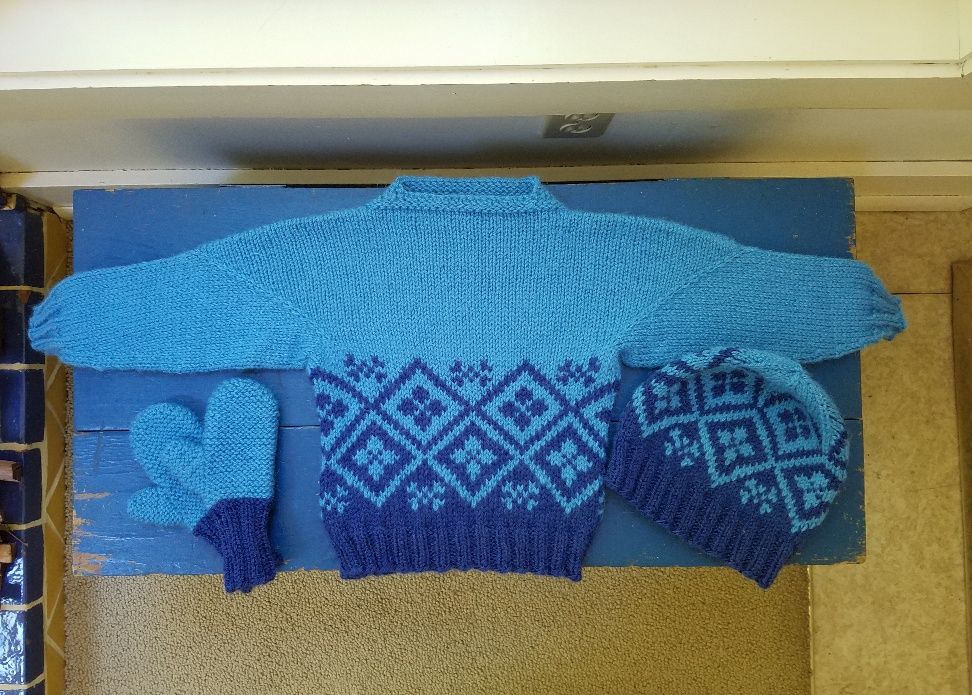A toddler-sized hand knitted sweater, hat, and mittens in stranded colorwork, using a large geometric motif that transitions from dark blue to light blue on the sweater and hat. Mittens have dark blue cuffs and light blue hands.