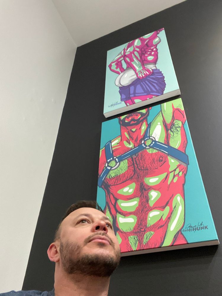 The picture shows the artist in front of two canvas art prints hanging on a customer’s wall. One artwork features a man getting ready for the gym, emphasizing his muscular glutes. The other depicts a man in a harness, with visible chest hair and armpits. Both pieces are bold and expressive, with vivid colors that stand out.