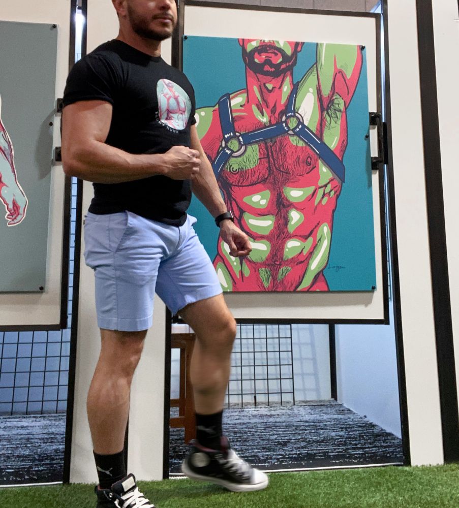 An illustration of a muscular man wearing a leather harness, his armpit exposed. The piece highlights sensuality and desire, created with strong lines and contrasting shadows. The artist stands in front of the art print