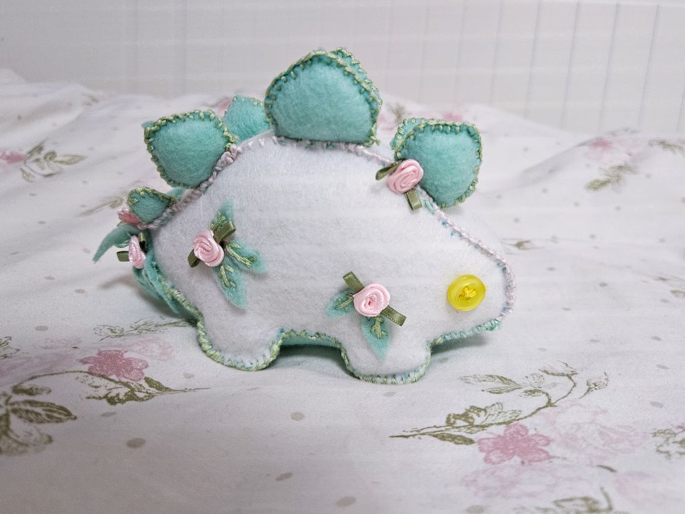 A stegosaurus plush, it's quite body, green plates and yellow eyes are light and the eyes are shiny.
The stegosaurus has roses and leaves as the patterns.
The stegosaurus is standing on a flower sheet that has pink flowers on it.